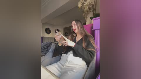 Media: Video of a young Asian woman with long brown hair in a black and white robe, sitting in a purple chair, using a curling iron on her hair.