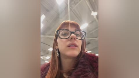 Media: A video of a fair-skinned woman with straight red hair, wearing black-rimmed glasses and a maroon fur coat, standing indoors with a high ceiling and visible lights.