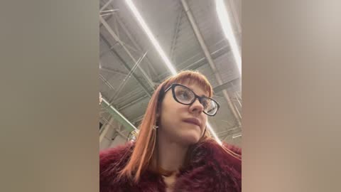 Media: A video of a young woman with long, straight, auburn hair and black-rimmed glasses, wearing a maroon fur coat, standing in a warehouse with exposed beams and industrial lighting.