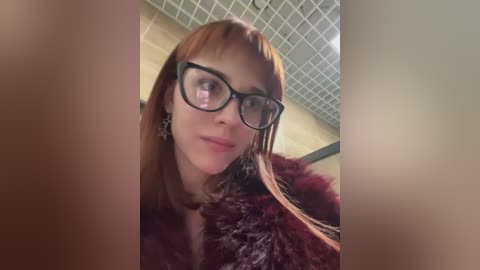 Media: Video of a woman with straight, reddish-brown hair, wearing black-framed glasses and a maroon fur coat, standing in a tiled bathroom with a grid ceiling.