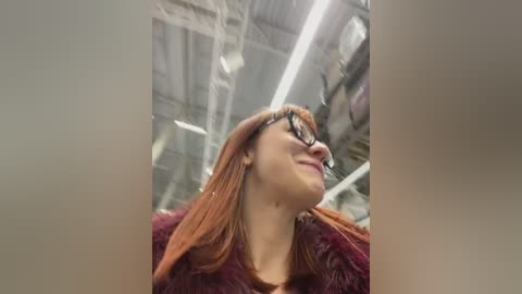 Media: A video of a smiling woman with long, straight, reddish-brown hair, wearing black-framed glasses and a burgundy fur coat, captured in an industrial setting with exposed metal beams and fluorescent lights.