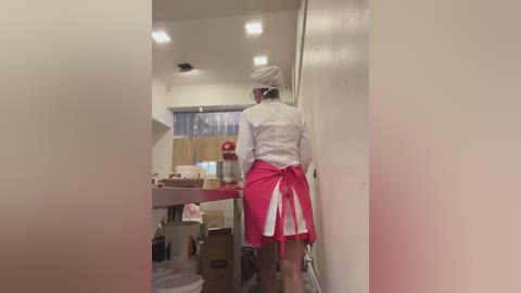 Media: Video of an elderly woman with white hair and glasses in a white lab coat and pink apron, standing in a narrow, sterile hallway with white walls and fluorescent lights, boxes and medical supplies on the floor.