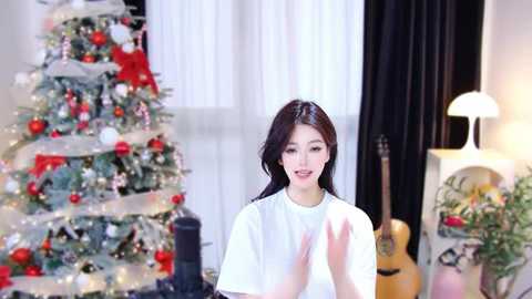 Media: A video of a smiling Asian woman with dark hair, wearing a white t-shirt, clapping in front of a decorated Christmas tree and guitar.