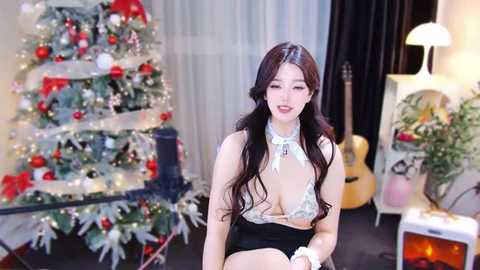 Media: Video of an East Asian woman with long black hair, fair skin, and a slender physique, wearing a low-cut, lacy white top and black shorts, smiling. Decorative Christmas tree and guitar in background.