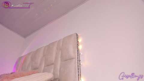 Media: A video of a modern bedroom featuring a beige, tufted headboard with string lights and a minimalist, white wall. The room has a cozy, soft ambiance.