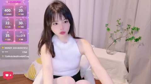 Media: A young Asian woman with long black hair, fair skin, and a slim physique, wears a white sleeveless top and black shorts. She sits on a yellow and white cushioned chair in a cozy room with a green plant on a table.