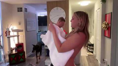 Media: Video of a topless blonde woman with medium breasts, holding a white towel, in a modern apartment. A Doberman dog and a man are present.