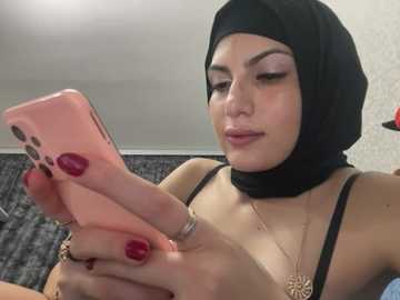 Media: A video of a light-skinned woman with dark hair in a black hijab, wearing a black tank top, holding a pink iPhone, with red-painted nails, against a beige background.