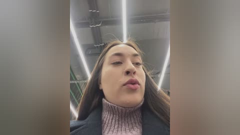 Video of a young woman with straight brown hair, light skin, and a light pink turtleneck sweater, puckering her lips, taken from a low angle, with a modern, industrial background featuring bright, linear lighting and metal structures.