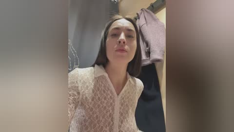 Media: A video of a young woman with straight, dark hair, wearing a white lace blouse, standing in a dimly lit bathroom with towels and a purple shirt hanging on hooks.