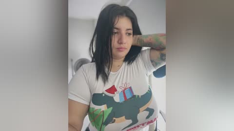 Media: Video of a young woman with shoulder-length black hair, wearing a white T-shirt with a cartoonish dinosaur and presents design, standing in a white-walled room. She has tattoos on her arm and a thoughtful expression.