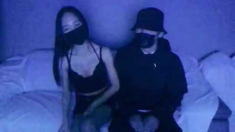 Media: Video of two Asian women in black face masks, one with long straight hair, the other with a bucket hat, seated on a bed in a dimly lit room with blue lighting.