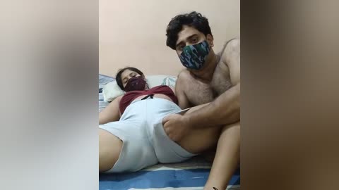 Media: Video of a shirtless South Asian man with a beard and dark hair, wearing a face mask, sitting on a bed next to a woman in a maroon top and light gray shorts.