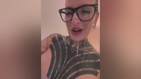 Media: Video of a woman with pale skin, black-framed glasses, and a large, intricate black tattoo on her chest, smiling broadly, her green eyes visible, in a dimly lit room.