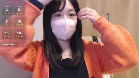 Media: Video of an East Asian woman in an orange sweater, wearing a mask, adjusting her hat indoors. Background shows a tiled wall and a digital temperature display.