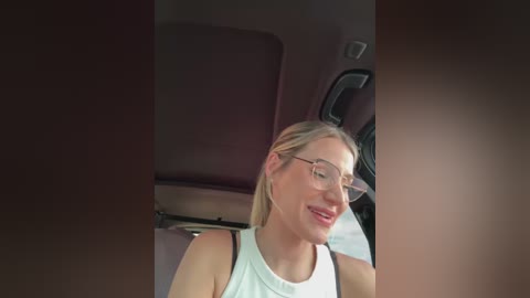 Media: A video of a smiling woman with blonde hair and glasses, wearing a white sleeveless top, seated in a car. The background shows the car's interior with a dark headrest and black leather seats.