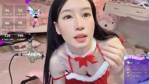 Media: Video of an East Asian woman with long black hair, wearing a revealing red and white Santa-themed lingerie, posing with a microphone in a cozy, pink-themed bedroom.