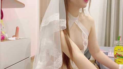 Media: A video shows an Asian woman with long brown hair in a white lace bridal gown, adjusting her veil in a room with pink walls, a white dresser, and a glass jar of eggs.