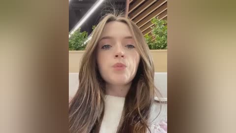 Media: A video of a young woman with long, light brown hair, fair skin, and pink lips, wearing a white top, standing in a modern indoor space with green plants and wooden panels in the background.