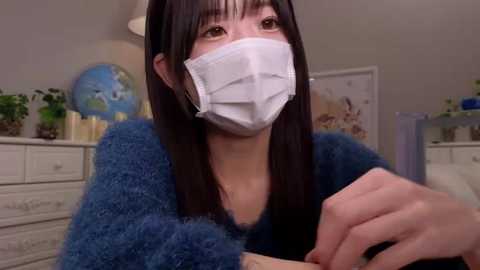 Media: Video of an East Asian woman with straight, dark hair and fair skin wearing a blue sweater, white mask, and a black shirt, sitting indoors with a globe and potted plants in the background.