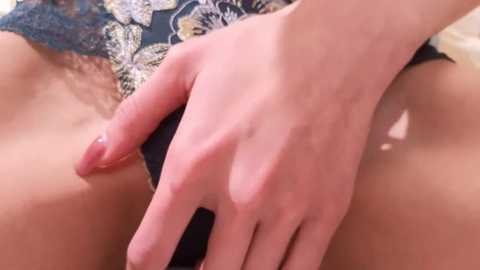 Media: Close-up video of a light-skinned hand gently pressing a black lace bra against a woman's chest, with a floral-patterned fabric in the background.