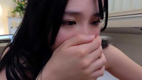 Media: A close-up video of a young Asian woman with long black hair, covering her mouth with her hands, in a modern kitchen setting.