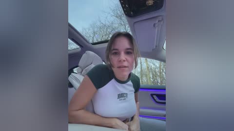 Media: Video of a Caucasian woman with light skin and shoulder-length brown hair, wearing a white and green short-sleeved shirt. She's leaning against the back seat of a car with a cloudy sky and bare trees visible outside.