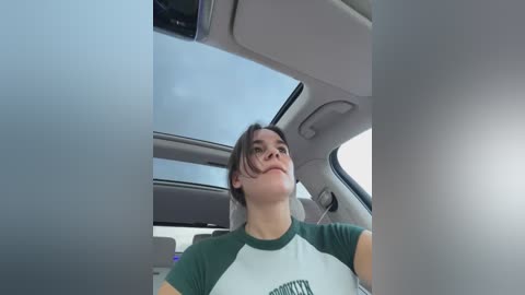 Media: Video of a young woman with straight, shoulder-length brown hair, wearing a green and white T-shirt, looking upward through the car's sunroof. The car's interior is modern with a gray ceiling and a partly visible steering wheel.