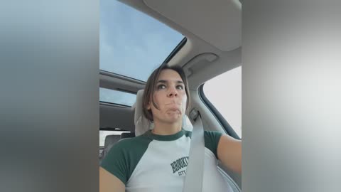 Media: A video captures a young woman with light skin and shoulder-length brown hair, wearing a white and green sports t-shirt, driving a car with a blue sky and a building visible through the window.