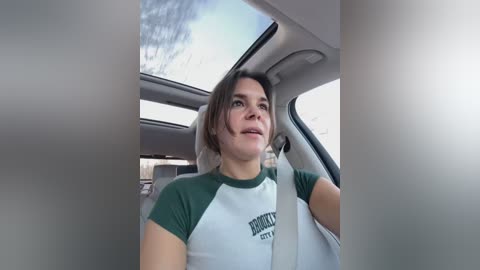 Media: Video of a young woman with shoulder-length brown hair, wearing a green and white athletic shirt, smiling while driving in a car.