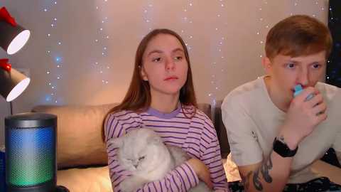 Media: Video of a young woman with long brown hair, wearing a striped shirt, holding a white cat, and a young man with short brown hair, both sitting on a beige sofa, surrounded by a cozy, dimly lit room with a blue LED lamp.