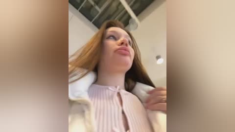 Media: Video of a young Caucasian woman with fair skin, long red hair, and a pink striped blouse, pouting, taken from a low angle, in a modern indoor setting with exposed ceiling beams and soft lighting.