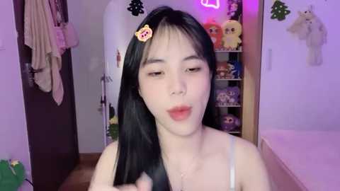 Media: Video of a young Asian woman with long black hair, wearing a white spaghetti-strap top, pouting with a \"LOL\" sticker on her forehead, in a cozy bedroom with plush toys and a bed.