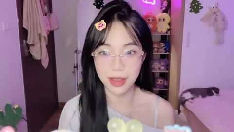 Media: Video of a young Asian woman with long black hair, wearing glasses and a white off-shoulder top, in a cozy room with plush toys and a bed.