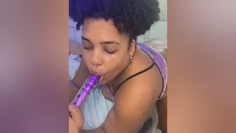 Media: Video of a young Black woman with natural curls, wearing a purple tank top, kneeling and licking a colorful dildo. She has a medium skin tone. Background shows tiled walls and a bed.