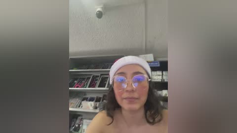 Media: Video of a young woman with light skin, wearing a Santa hat and round glasses, standing in a brightly lit, messy room filled with shelves of books and electronic devices.