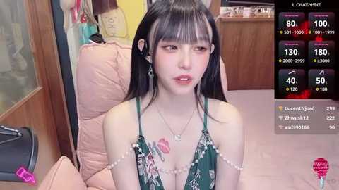 Media: Video of an East Asian woman with long black hair, wearing a green floral dress, sitting on a pink couch in a dimly lit room.