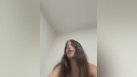 Media: A blurred, motion-blurred video of a young woman with long brown hair, standing in a white-walled room with a corner ceiling. Her face is partially visible, with a slight smile.