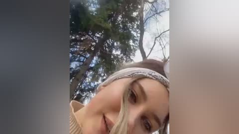 Media: Video of a young woman with light skin and long blonde hair partially covered by a headband, smiling. She's outdoors with tall, leafless trees and a cloudy sky in the background.