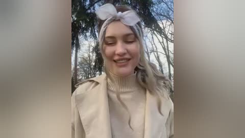 Media: Video of a smiling young woman with fair skin, long blonde hair, and a white headband, wearing a beige trench coat, set against a blurred forest background.