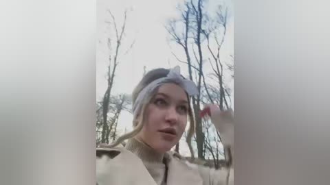 Media: Video of a young woman with light skin, wearing a white headband and beige coat, looking contemplative outdoors in a winter forest.