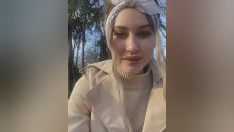 Media: Video of a smiling Caucasian woman with light skin, wearing a beige coat and headband, standing outdoors in a forest with tall trees and blue sky in the background.