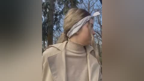 Media: Video of a woman with long blonde hair, wearing a beige trench coat and a bandana, standing in a forest.