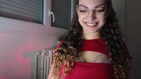 Media: Video of a smiling woman with curly hair, glasses, and a red shirt, revealing a pink bra underneath, in a dimly lit room with a window and radiator.