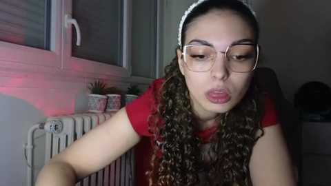 Media: A video of a young woman with light skin, long curly brown hair, and glasses, wearing a red shirt, leaning over a white radiator in a dimly lit room.