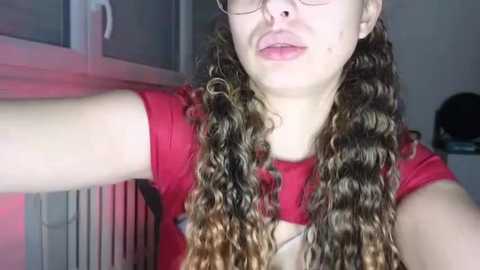 Media: Video of a fair-skinned woman with long, curly, brown hair and glasses, wearing a red t-shirt. She has acne on her cheeks and is taking a selfie.