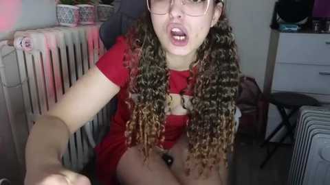 Media: A video of a young woman with curly, multi-colored hair, wearing a red top and glasses, sitting on a chair in a cluttered room, holding a selfie stick.