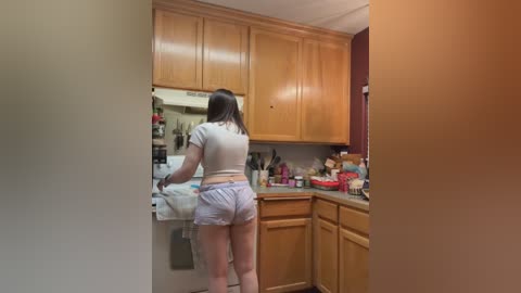 Media: Video of a woman with straight, shoulder-length brown hair, wearing a white crop top and light blue shorts, cooking in a modern, wooden kitchen with cabinets and countertop cluttered with kitchen items.