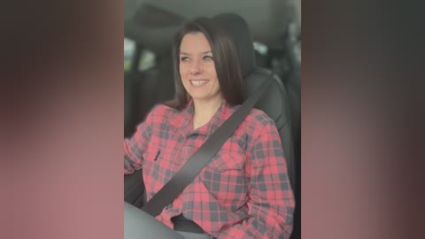 Media: A video of a smiling Caucasian woman with long dark hair, wearing a red and black plaid shirt, seated in a car with a seatbelt on, with blurred background.