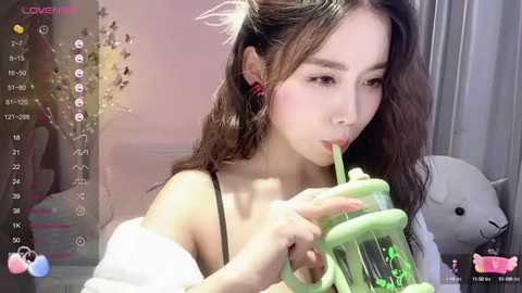 Media: Video of an Asian woman with long brown hair, wearing a black spaghetti strap top, drinking from a green straw, in a softly lit room with a pink wall and a teddy bear.